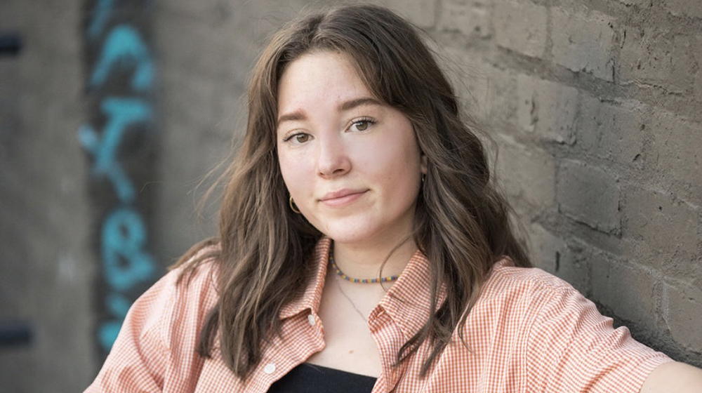 Portrait of a current Gannon Scholar with shoulder-length brown hair, wearing an orange checkered shirt over a black top and a beaded necklace, leaning against a brick wall with graffiti in the background.